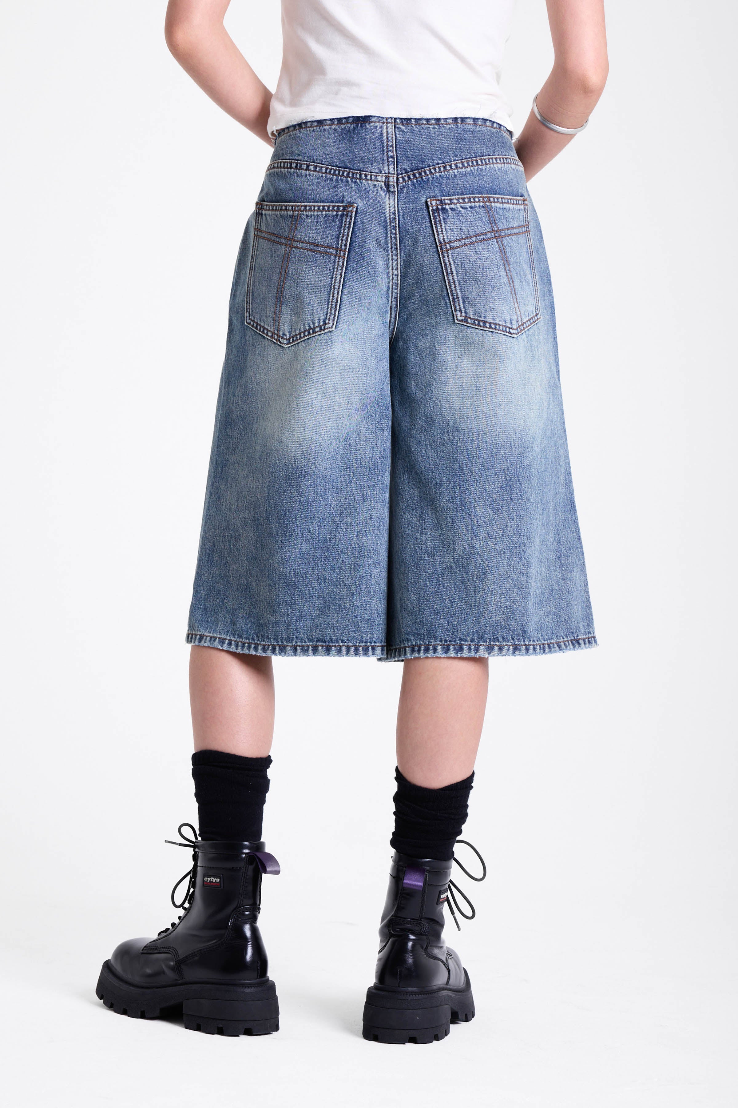 Mid blue denim longline jorts with no waistband and silver piercing hardware detail, worn by model