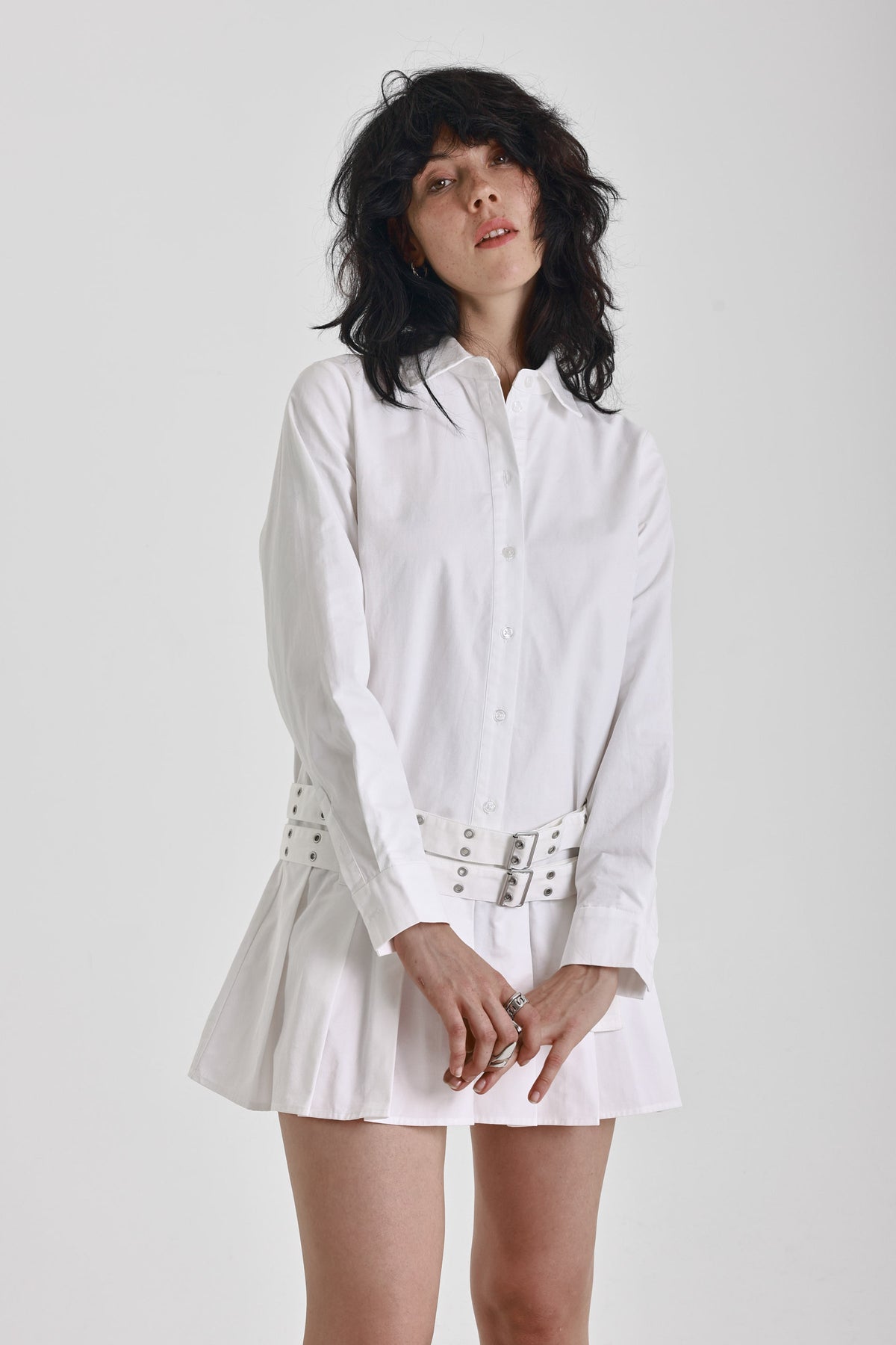 White mini shirt dress with long sleeves, pleated skirt and double eyelet belt, worn by model