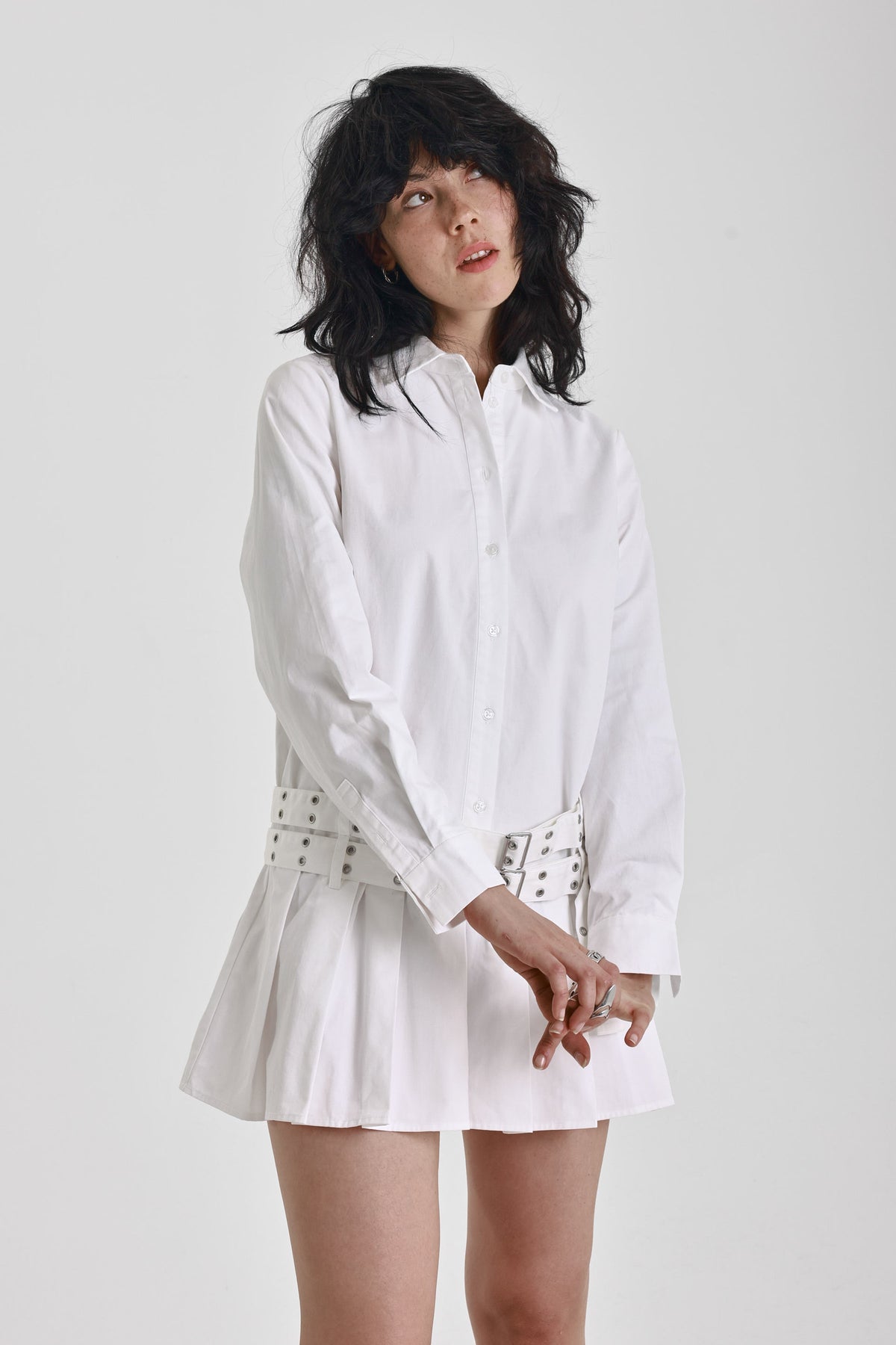 White mini shirt dress with long sleeves, pleated skirt and double eyelet belt, worn by model