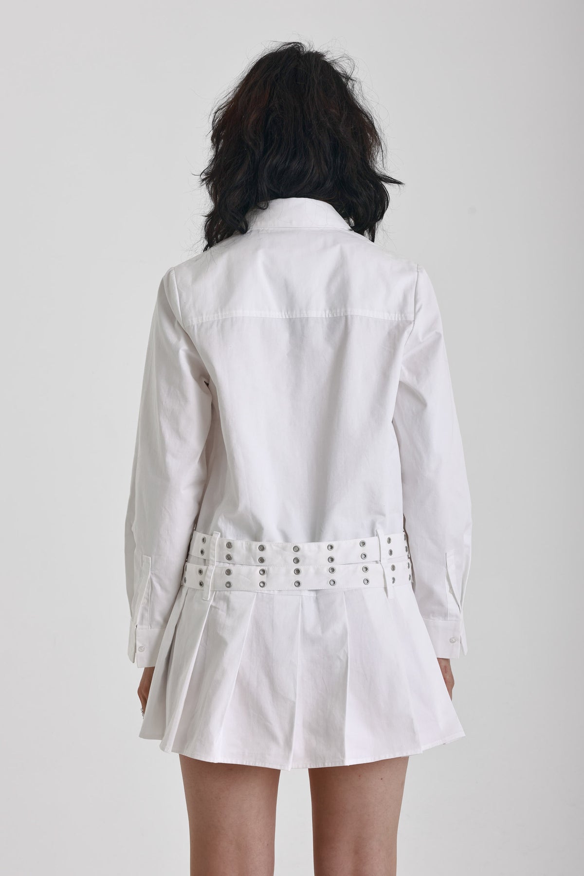 White mini shirt dress with long sleeves, pleated skirt and double eyelet belt, worn by model