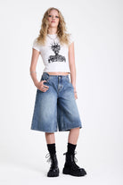Mid blue denim longline jorts with no waistband and silver piercing hardware detail, worn by model