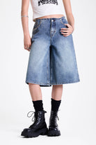Mid blue denim longline jorts with no waistband and silver piercing hardware detail, worn by model