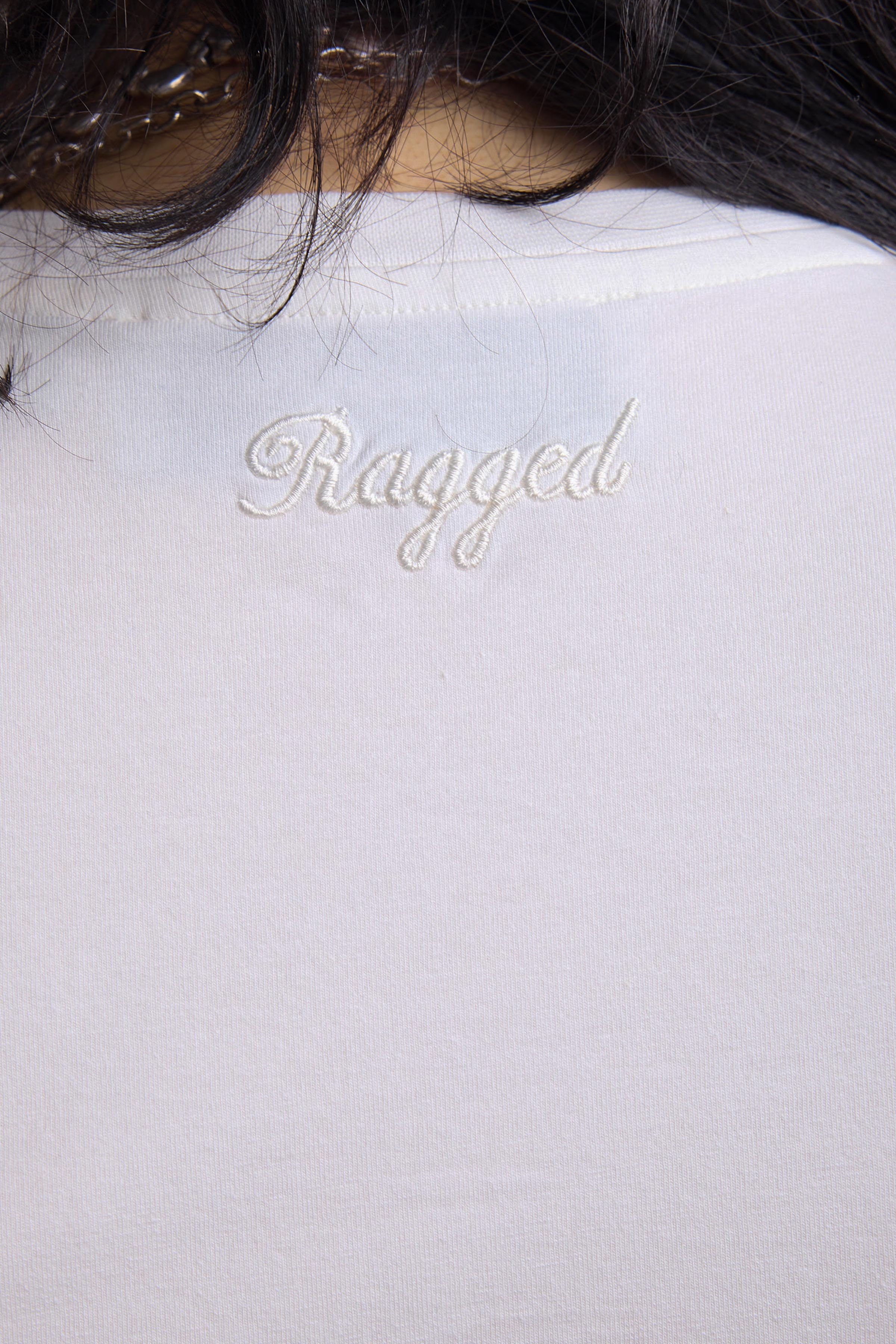 Close up of embroidered Ragged logo on white shrunken baby t-shirt, worn by a model