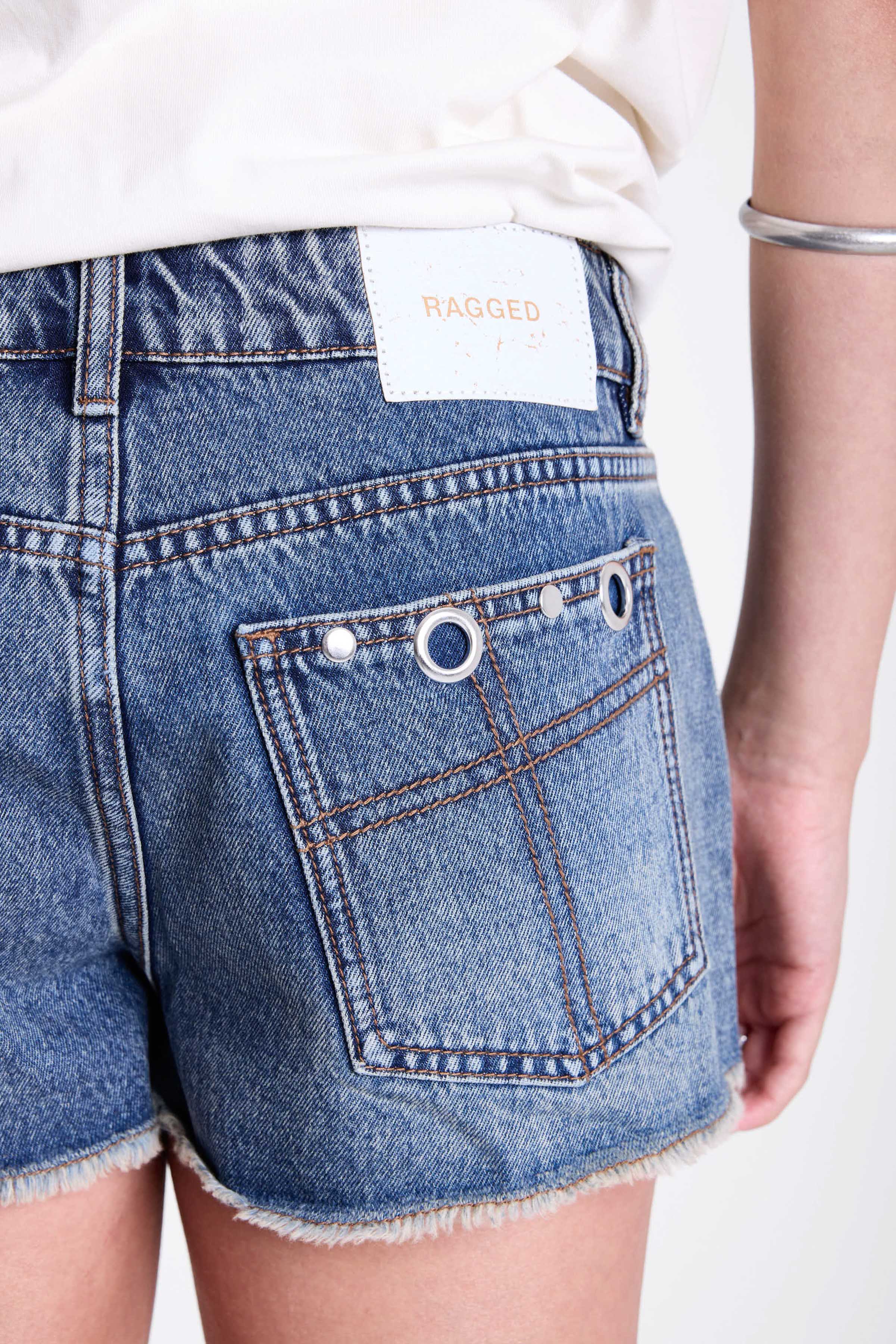 Mid blue denim mini shorts with raw hem and silver eyelet rivet and stud detail, worn by model