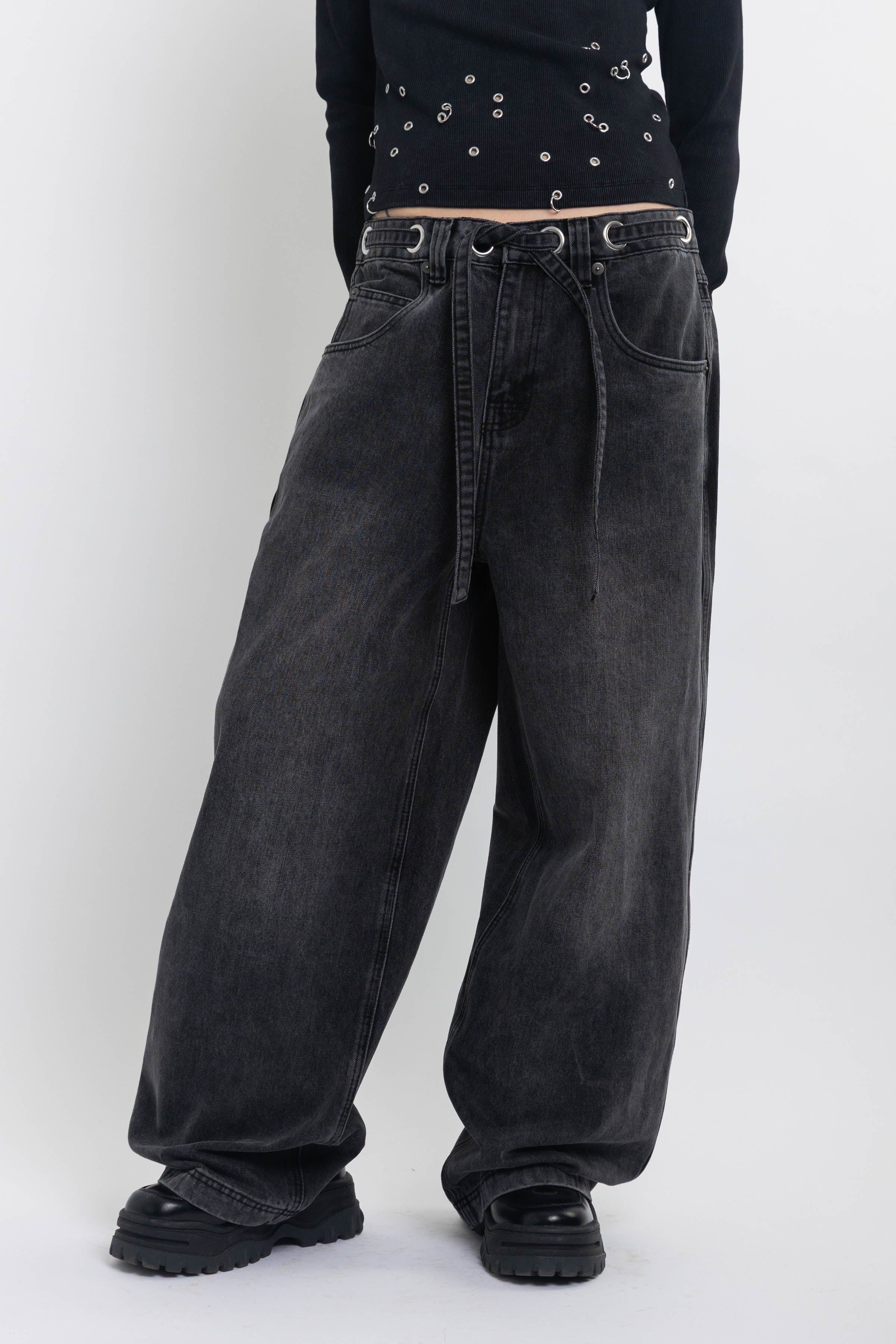 Washed black ultra baggy colossus jeans with silver eyelet waistband detail, worn by model 