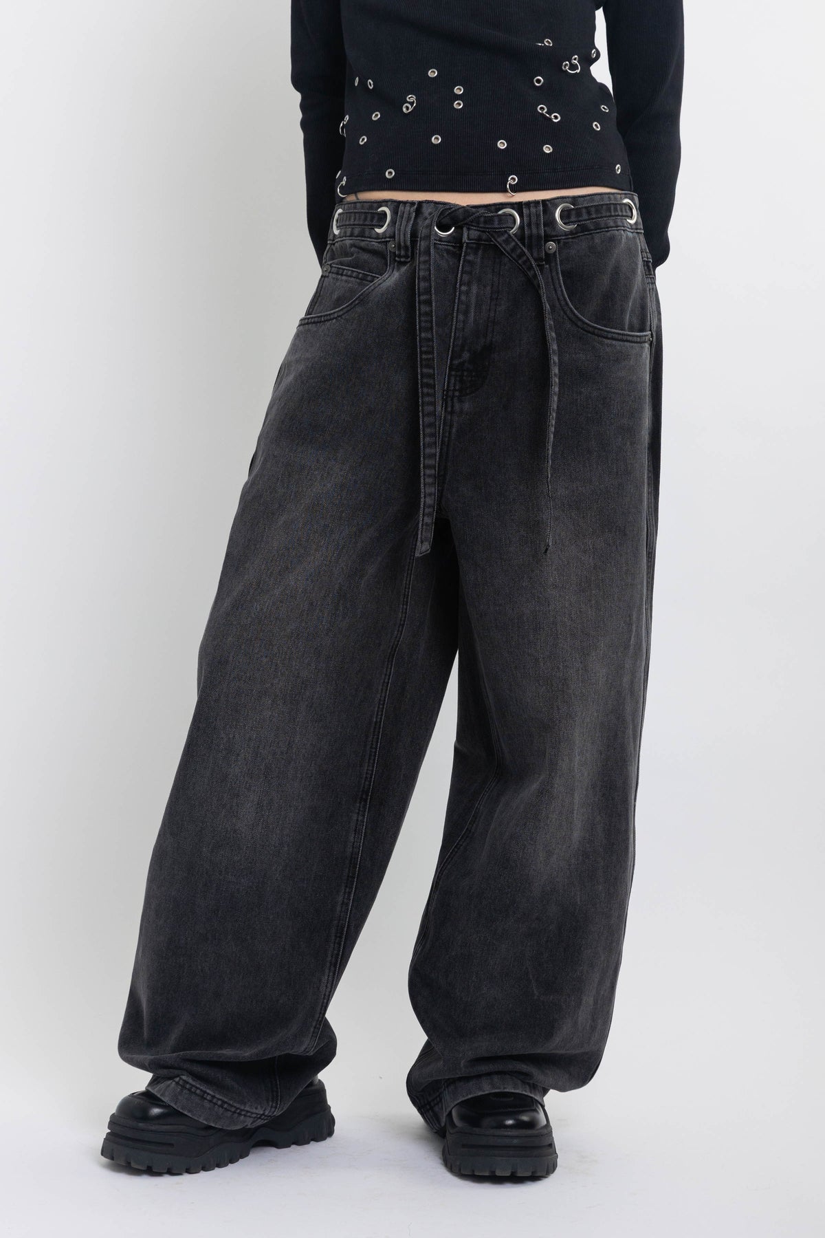 Washed black ultra baggy colossus jeans with silver eyelet waistband detail, worn by model 