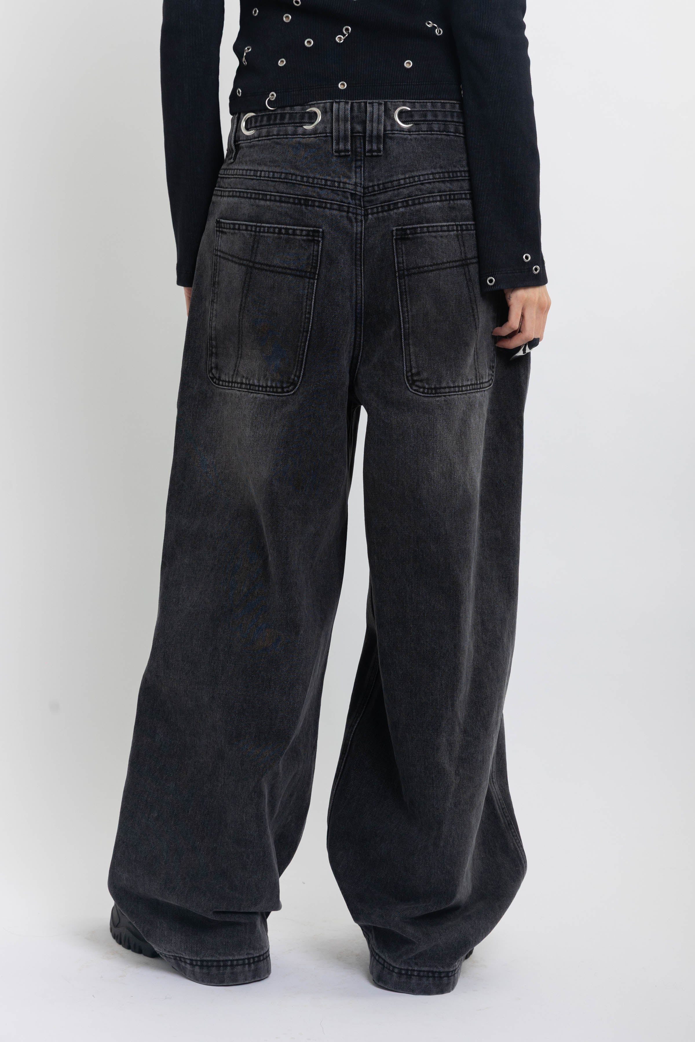 Washed black ultra baggy colossus jeans with silver eyelet waistband detail, worn by model 