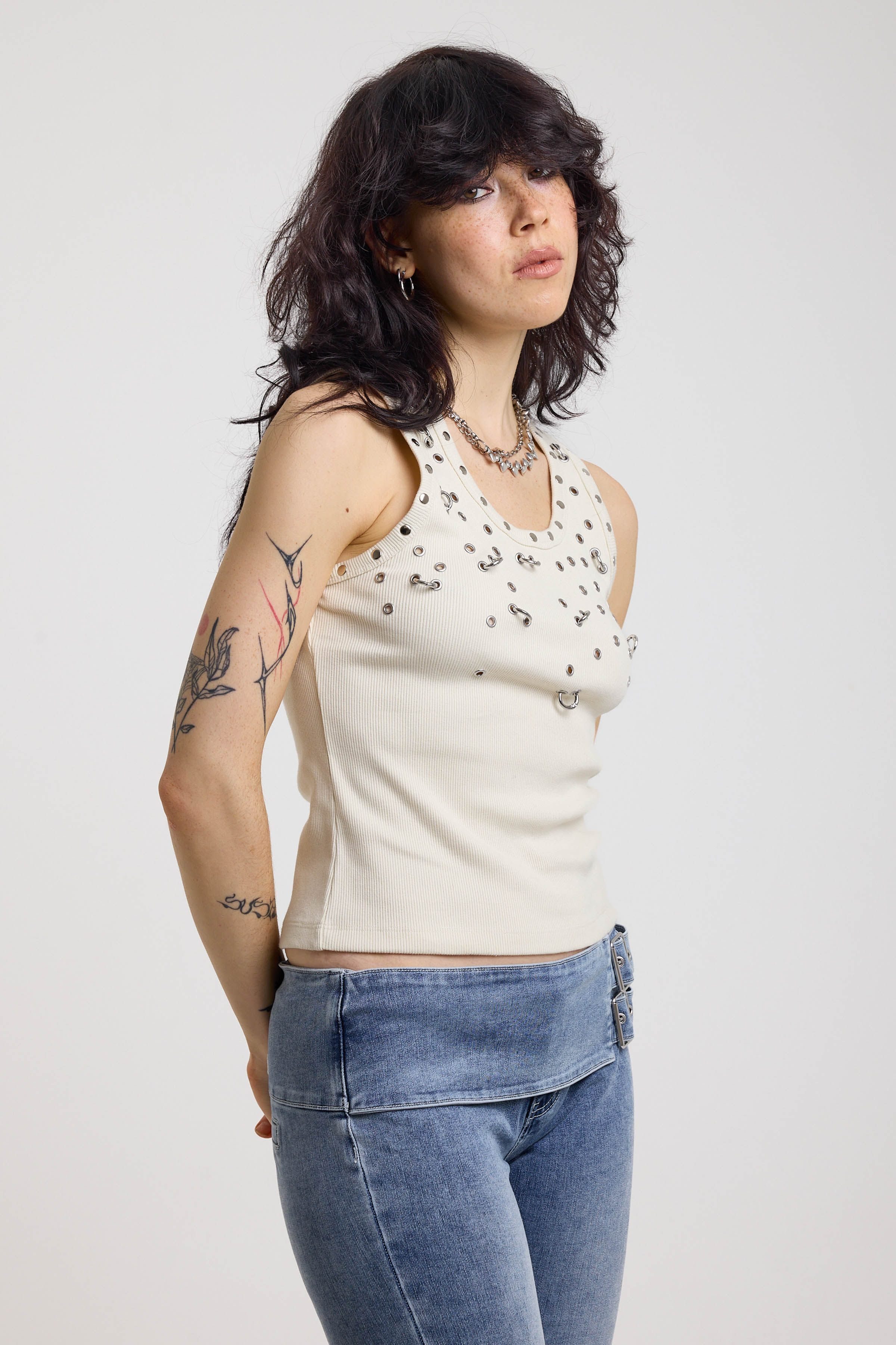 Ecru ribbed tank top with piercing and stud hardware details, worn by a model