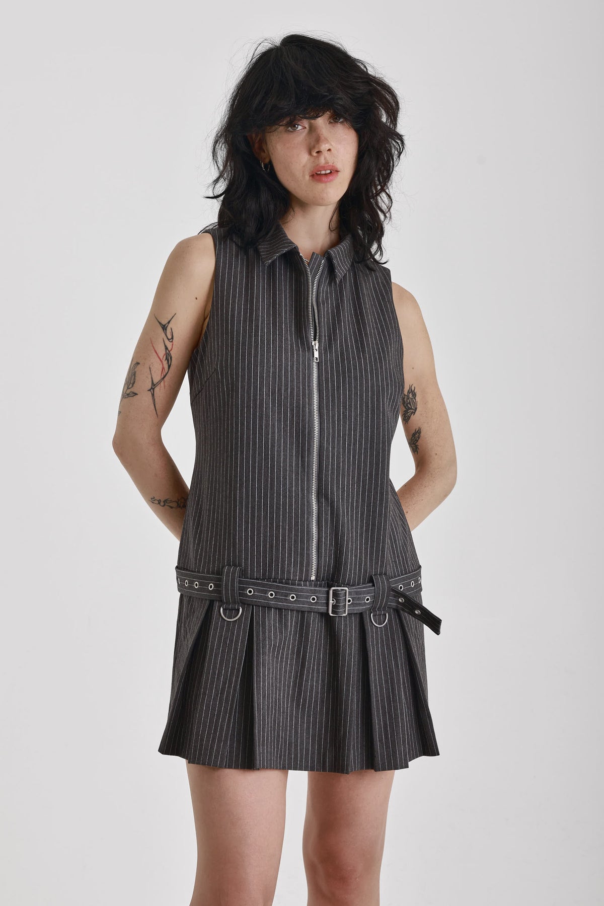 Grey pinstripe zip through mini dress with pleated skirt and silver eyelet belt and D-ring hardware, worn by model